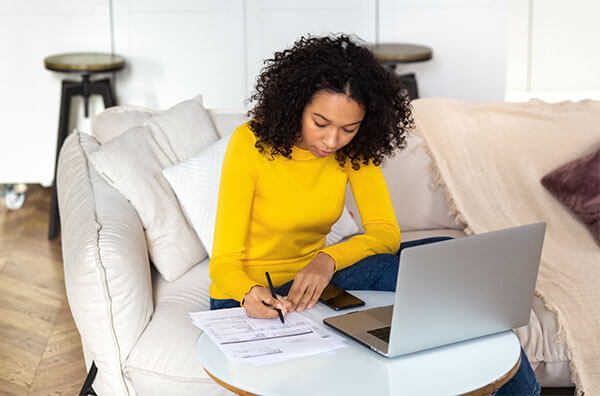 Woman sitting on couch while leaning over table to write on document with phone and laptop resting next to her.