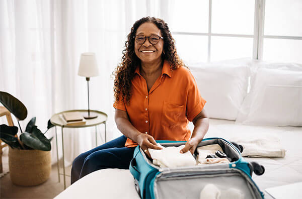 Happy lady smiling while packing suitcase resting on bed with large windows, bed side table and plant in background.