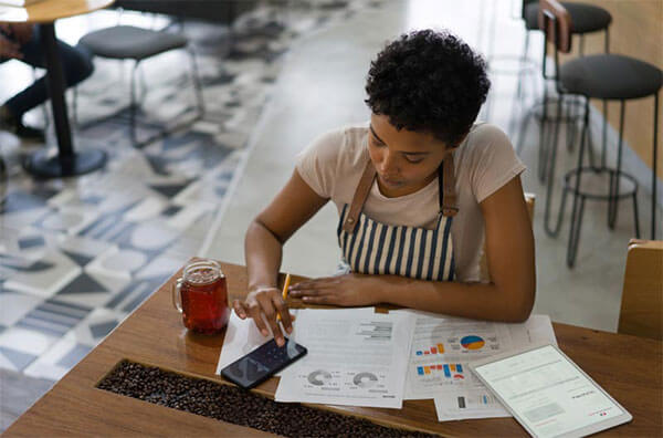 Woman sitting at cafe table wearing apron using phone to calculate charts resting on table in front of her.