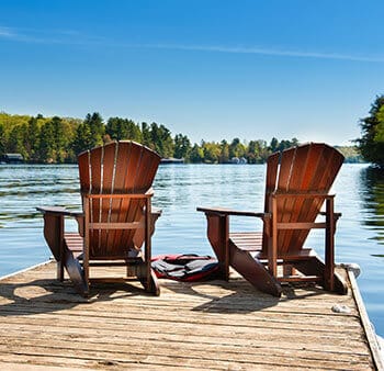 Two Adirondack chairs on the edge of a dock facing the lake, large trees, and big blue sky in front of them.