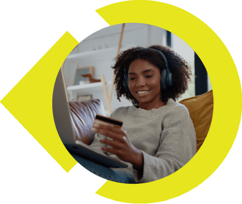Woman laying on couch with headphones on, laptop propped up on her legs, and credit card in hand.