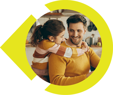 Father sitting at kitchen table with young daughter hugging him around the neck while they smile at each other.