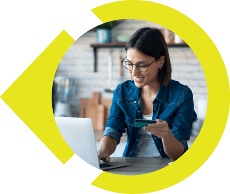 Woman with glasses and jean shirt sitting at kitchen table holding credit card in one hand and typing on laptop with the other.