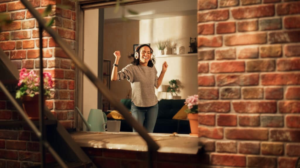 A woman wearing headphones dances joyfully inside her home, seen through a brick-framed window, with potted flowers on the windowsill. The atmosphere is lively.