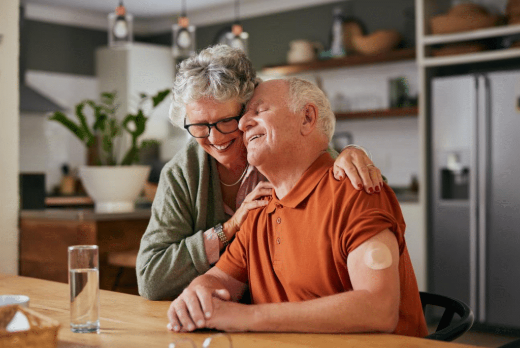 Elderly couple sharing a joyful hug at a kitchen table. The man, wearing an orange shirt, has a medical patch on his arm. The atmosphere is warm and loving.