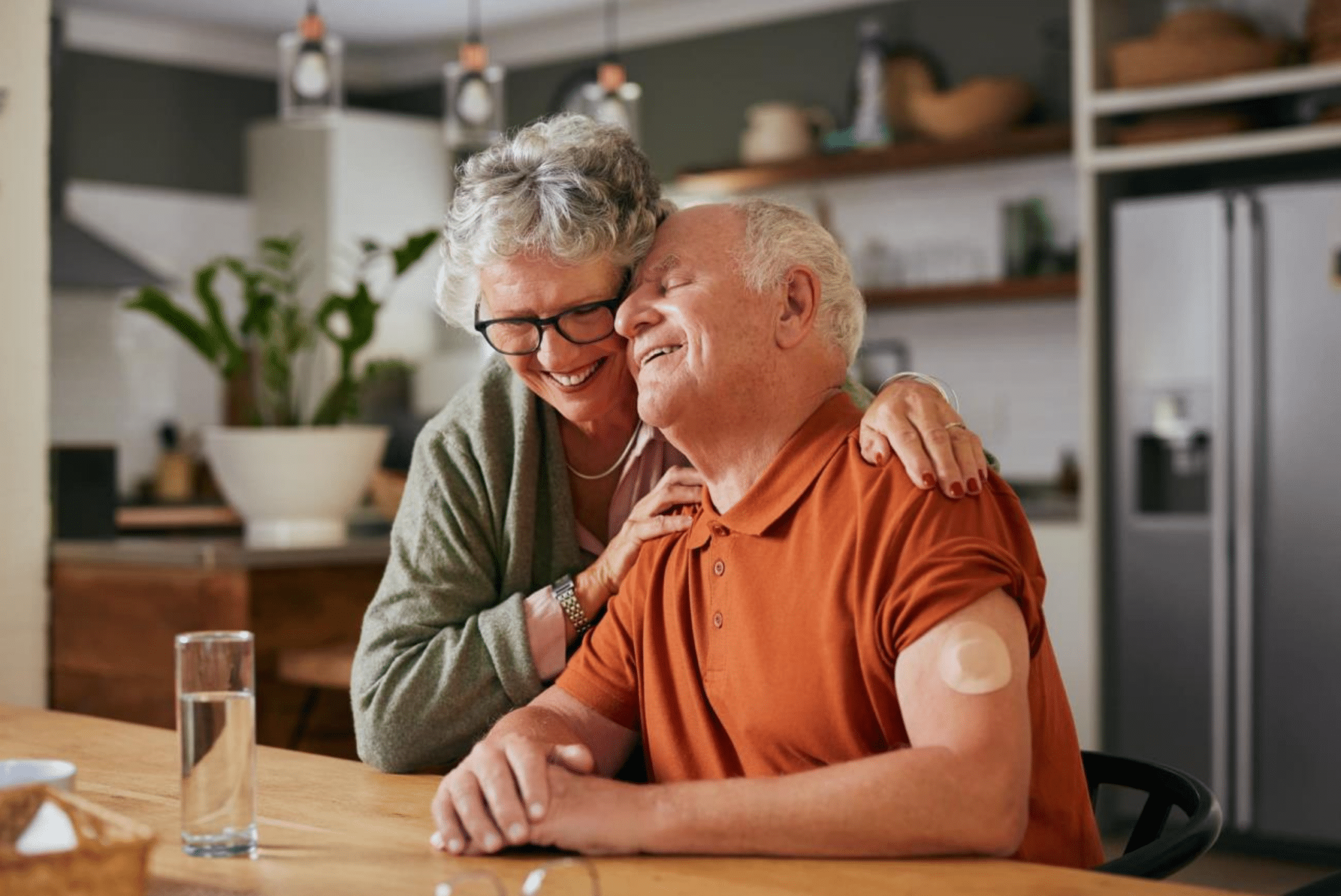 Elderly couple sharing a joyful hug at a kitchen table. The man, wearing an orange shirt, has a medical patch on his arm. The atmosphere is warm and loving.
