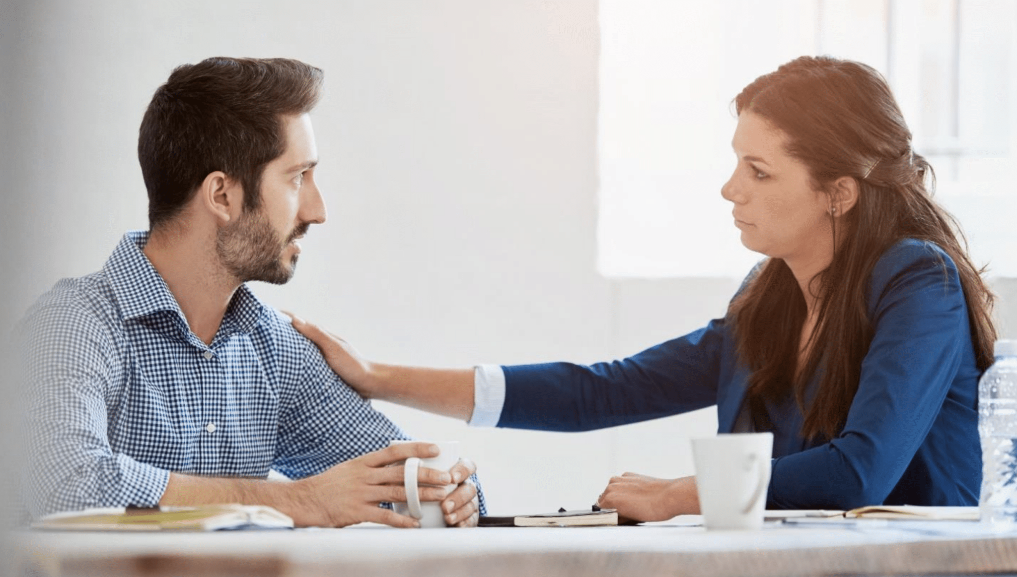 A woman in a blue blazer gently rests her hand on a man's shoulder, offering comfort. They sit at a table with coffee mugs, creating a supportive atmosphere.